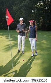 Nice Middle Aged Couple Playing Golf During A Sunny Day. They Both Reach The Green Of The Fifteenth Hole And Its Red Flag And Are Smiling At Each Other. They Are Wearing Caps And Sportswear Outfits