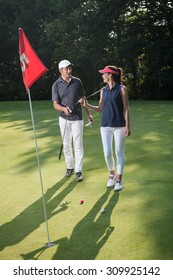 Nice Middle Aged Couple Playing Golf During A Sunny Day. They Both Reach The Green Of The Fifth Hole And Its Red Flag And Are Smiling At Each Other. They Are Wearing Caps And Sportswear Outfits