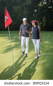 Nice Middle Aged Couple Playing Golf During A Sunny Day. They Both Reach The Green Of The Fifth Hole And Its Red Flag And Are Smiling At Each Other. They Are Wearing Caps And Sportswear Outfits