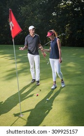 Nice Middle Aged Couple Playing Golf During A Sunny Day. They Both Reach The Green Of The Fifth Hole And Its Red Flag And Are Smiling At Each Other. They Are Wearing Caps And Sportswear Outfits