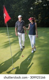 Nice Middle Aged Couple Playing Golf During A Sunny Day. They Both Reach The Green Of The Fifth Hole And Its Red Flag And Are Smiling At Each Other. They Are Wearing Caps And Sportswear Outfits