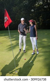 Nice Middle Aged Couple Playing Golf During A Sunny Day. They Both Reach The Green Of The Fifth Hole And Its Red Flag And Are Smiling At Each Other. They Are Wearing Caps And Sportswear Outfits