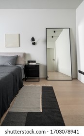 Nice modern bedroom with white walls with a relief picture and a parquet with a multicolored carpet. There is a bed with gray linens and pillows, dark lamps, black nightstand, tall floor mirror. - Shutterstock ID 1703078941