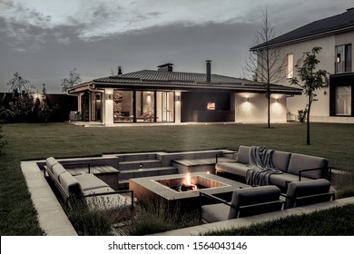 Nice modern estate with a two storey house, spa building, green lawn with trees and a relax zone with a burning fire pit on the background of the cloudy sky. Lamps are luminous. Horizontal.