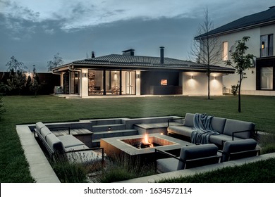 Nice Modern Estate With A Two Storey House, Spa Building, Green Lawn With Trees And A Relax Zone With A Burning Fire Pit On The Background Of The Cloudy Sky. Lamps Are Luminous. Horizontal.