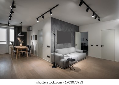 Nice modern interior with different walls and a parquet. There is a wooden table with chairs, windows with curtain, luminous lamps, doors, gray sofa with a laptop, black round stand, conditioner.