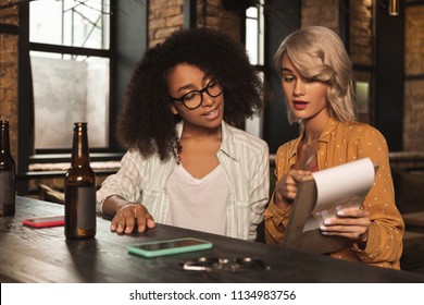 Nice Offer. Pleasant Young Women Sitting At The Bar Counter At The Pub And Checking Its Menu, Deciding Upon Their Order
