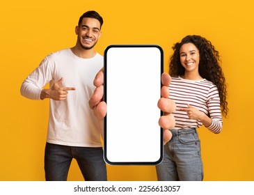 Nice Offer. Smiling Middle Eastern Couple Pointing At Big Blank Smartphone In Hands, Arab Man And Woman Demonstrating Copy Space For Mobile Advertisement, Standing On Yellow Background, Mockup - Shutterstock ID 2256613057