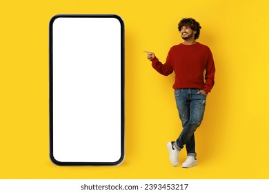 Nice online offer. Positive cheerful casual handsome young indian guy pointing at big smartphone with white blank screen, isolated on yellow studio background, mockup, copy space