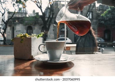 Nice Picture Of Someone With A Glass Carafe Pouring Black Coffee Into A White Porcelain Coffee Cup Next To A Green Plant On A Concrete Plateau On A Wooden Table In Front Of A Stained Glass Window.