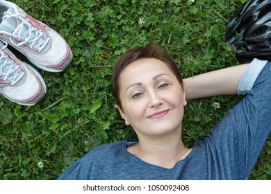 Nice Portrait Of Middle Aged 40 50 Years Old Woman Lying Down On A Grass Lawn.  Sneakers And A Bike Helmet Are Nearby. Top View, Close-up Face.