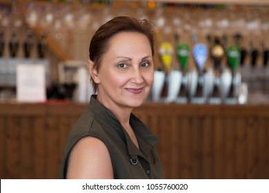 Nice Portrait Of A Middle Aged Older Woman In A Pub. Professional Headshot Of 45 50 Year Old Relaxed Female Wearing A Sleeveless Khaki Top, Looking At Camera And Smiling. Lifestyle Shot.