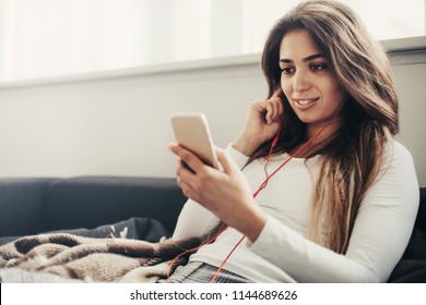 Nice and positive girl sit and hold phone in hands. Also woman holds finger on one of headphones. She is listening to music. Her legs are covered with blanket - Shutterstock ID 1144689626