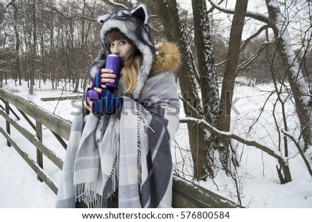 Nice pretty cheerful girl  in the wolf hood and fur scarf in the form of paws and with fingerless gloves, wrapped a blanket, holding cup of hot tea in a knit cover, handmade