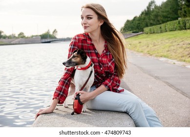 Nice pretty long haired girl in tartan red shirt sit at the river embankment with smiling fox-terrier.  Look left. 