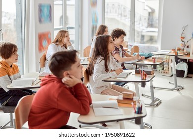 Nice Pupils Are Listening Carefully To Their Tutor. Elementary School Kids Sitting On Desks And Reading Books In Classroom