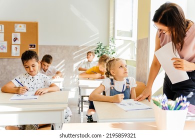 Nice Pupils Are Listening Carefully To Their Tutor. Primary School Kids Sitting On Desks And Working In Classroom.