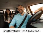 Nice Ride. Portrait of happy male driver riding car looking at cheerful beautiful lady sitting inside auto on back passenger seat, female using cell phone and talking with guy, windshield view