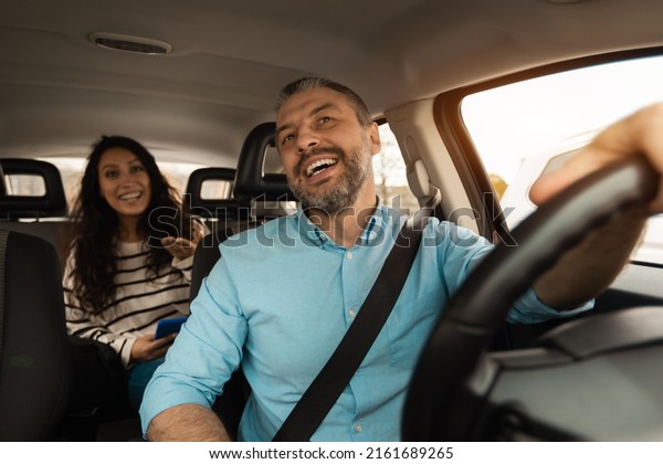 Nice Ride. Portrait of happy male driver riding car\
looking at cheerful beautiful lady sitting inside auto on back\
passenger seat, female using cell phone and talking with guy,\
windshield view