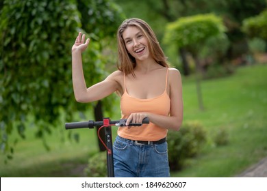 Nice To See You. Long-haired Cute Girl Riding Scooter In The Park And Raising Her Hand