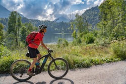 Nice Senior Woman Riding Her Electric Mountain Bike Above The Freiberg Lake And A Big Ski Flying Hill In The Allgau Alps Near Oberstdorf, Bavaria, Germany