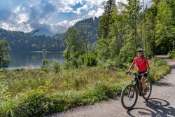 Nice Senior Woman Riding Her Electric Mountain Bike Above The Freiberg Lake And A Big Ski Flying Hill In The Allgau Alps Near Oberstdorf, Bavaria, Germany
