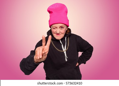 Nice Shot Of Gangsta Granny. An Old Woman Went Off The Rails And Dressed In Hip-hop Style, Her Mood Is Uplifted: She Winks And Shows A Peace Sign. Radial Pink To White Gradient In The Background.