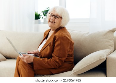 A Nice, Sweet Old Lady In A Brown Pantsuit Is Sitting On The Couch Holding A Laptop On Her Lap And Looking At The Camera With A Slight Smile On Her Face