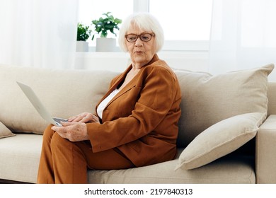 A Nice, Sweet Old Lady In A Brown Pantsuit Is Sitting On The Couch Holding A Laptop On Her Lap And Looking At The Camera With A Slight Smile On Her Face