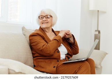 A Nice, Sweet Old Lady In A Stylish Jacket And Pants Is Sitting On A Beige Sofa With A Laptop, Smiling Pleasantly At The Camera And Folding Her Hands Near Her Face