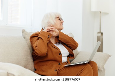 A Nice, Sweet Old Lady In A Stylish Jacket And Pants Is Sitting On A Beige Sofa With A Laptop, Smiling Pleasantly At The Camera And Folding Her Hands Near Her Face