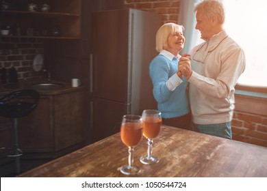 Nice And Warm Picture Of Old Couple Dancing Together In The Kitchen. They Have Left Two Full Wineglasses Of Wine On The Table. They Are Enjoying Company Of Each Other.