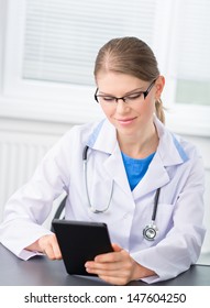 Nice Woman Doctor Sitting At The Desk And Pointing On Tablet PC. Beautiful Blond Caucasian Medical Female With Stethoscope, Ipad Wearing Eyeglasses In Hospital.    