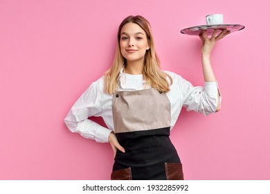 nice woman waitress in apron, offering cup of delicious tasty coffee on tray, stand smiling, friendly staff of restaurant. isolated over pink studio background