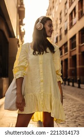 Nice Young Caucasian Brunette Woman With Headphones Spends Leisure Time In City Sunny Weather. Model Wears Yellow Summer Dress, Bag. Lifestyle Concept