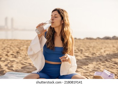 Nice young caucasian girl, closing eyes, quenches thirst by drinking water sitting on sand by sea. Blonde wears blue top, shorts and white shirt in summer. Weekend concept, leisure