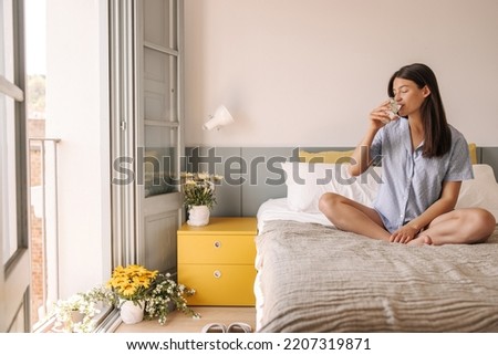 Nice young caucasian girl drinks water, crossing legs sitting on bed in room. Woman with brunette hair closes her eyes in pleasure. Good morning concept