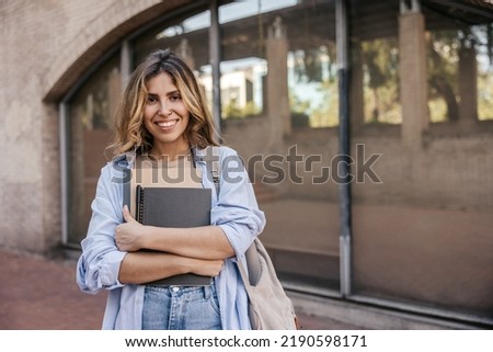Nice young caucasian girl with study notebooks, looking at camera standing on street. Blonde woman wears casual clothes in spring. Education concept