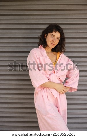 Nice young caucasian woman looks into camera with her arms around waist standing near rolling wall. Brunette wears pink summer shirt on casual day. Relaxed lifestyle, concept
