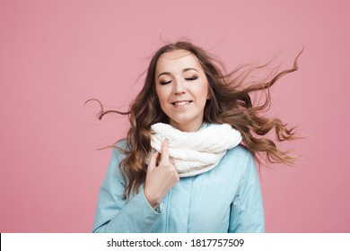 Nice Young Woman In A Blue Winter Jacket And Scarf, Her Hair In The Wind. Portrait On A Pink Background