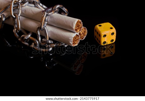 Nicotine and\
gambling addiction. Three cigarettes, rewound by a chain and a dice\
on a black background with\
reflection