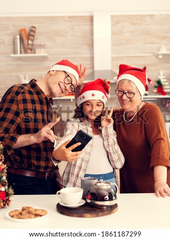 Niece taking selfie with granddparents celebrating christmas in decorated kitchen. Happy cheerful multi generation family using phone to take photo wearing santa hat doing hand gesture with xmastree