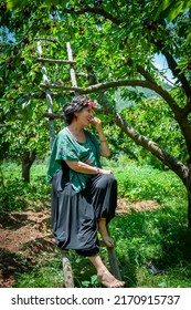 Nif, Arpacık, Muğla, 20 June 2022 - Unidentified Woman Wearing A Pretty Flower Crown And Decorative Cherry Top Sitting On A Wooden Ladder Eating A Cherry Picked From A Tree In A Cherry Orchard