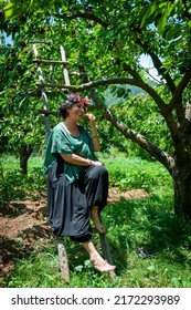 Nif, Arpacık, Muğla, 20 June 2022 - Unidentified Woman Wearing A Pretty Flower Crown And Decorative Cherry Top Sitting On A Wooden Ladder Eating A Cherry Picked From A Tree In A Cherry Orchard