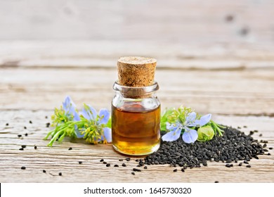 Nigella Sativa Oil In A Bottle, Seeds And Twigs Of Black Caraway Seeds With Blue Flowers And Green Leaves Of Kalingi On A Wooden Board Background