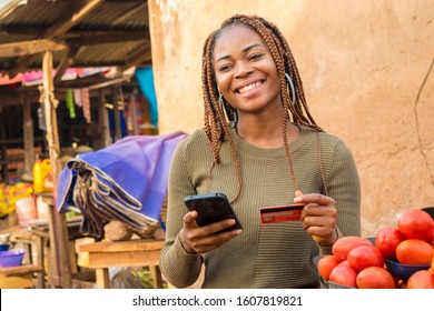 Nigerian Lady Selling In A Local Nigerian Market Using Her Mobile Phone And Credit Card To Do A Transaction Online Smiling