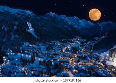 Night Aerial View Of Madonna Di Campiglio City, Italy On Christmas Night. Alpine Town In The Snow. Large Yellow Moon In The Starry Sky In The Alps. Ski Town At Night Lights.