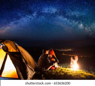 Night Camping. Charming Pair Tourists Sitting And Kissing Near A Campfire And Tent Under The Stars. On The Background Luminous Village In The Valley. Astrophotography