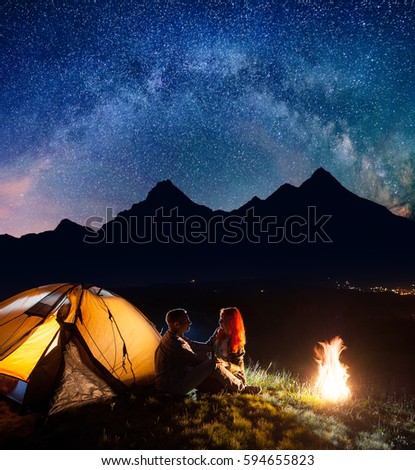 Night camping. Happy couple hikers sitting face to face in front tent near campfire under shines starry sky. On the background silhouettes of the high mountains and luminous village in the valley