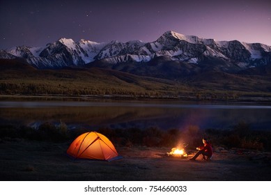 Night Camping. Man Tourists Sitting In The Illuminated Tent Near Campfire Under Amazing Sunset Evening Sky In A Mountains Area. Snow Mountain In The Background